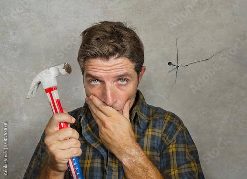 funny portrait of man holding hammer after driving a nail for hanging a frame but making a mess cracking the wall as a disaster DIY guy and messy domestic repair task photo