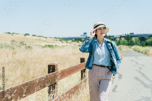 Beautiful asian chinese young woman tourist walk in grasslands. charming traveler in hat relax enjoy sunshine outdoors on mountain beside wooden fence with blue sky. smiling girl sightseeing nature © PR Image Factory