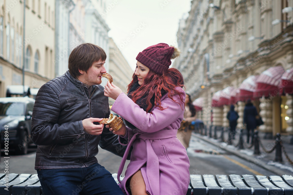 Fototapeta premium Portrait of beautiful young couple eating Trdelník in the street. Having fun in winter of the european city. Vacation and holiday concept