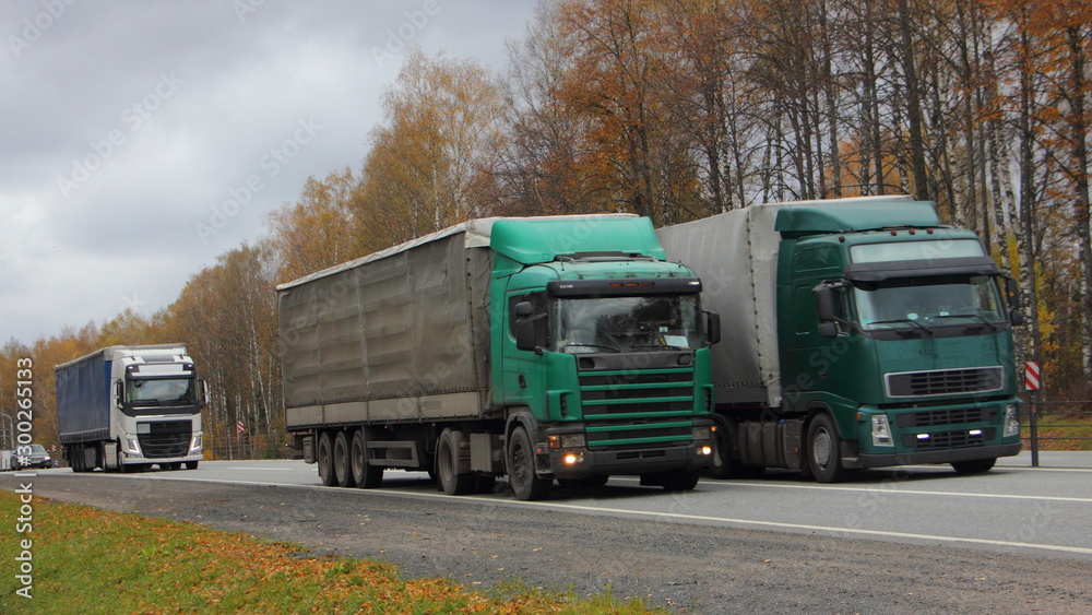 Three European green semi trucks with awning trailers overtaking on asphalt country highway on autumn day, front side view, transport logistics, goods delivery, beautiful road landscape