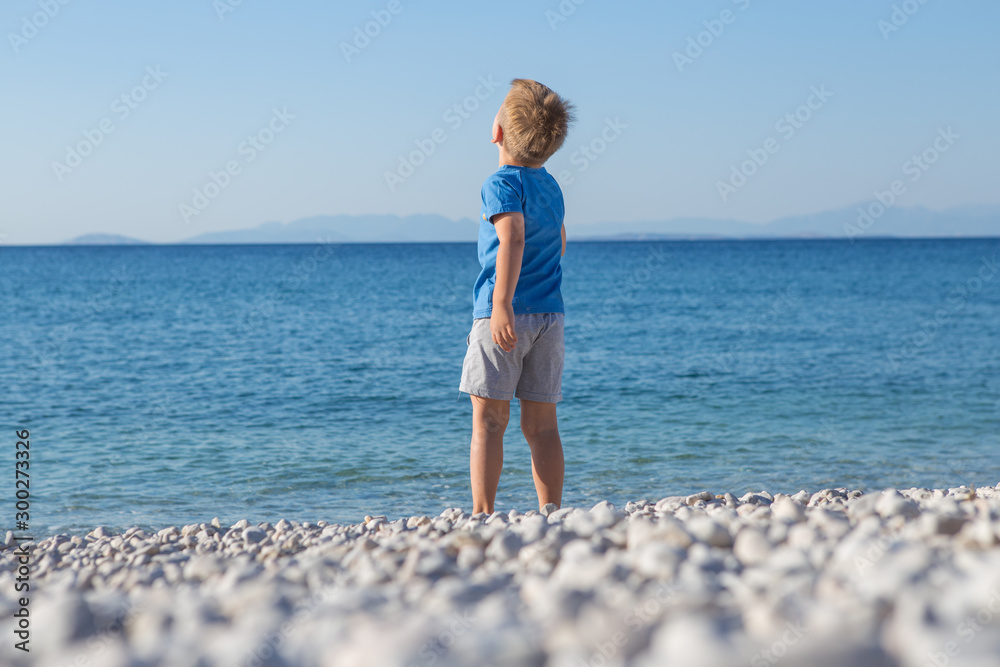 Cute European little boy in a blue swimming T-short is standing in the sea. He is enjoying his summer holidays. Back view.