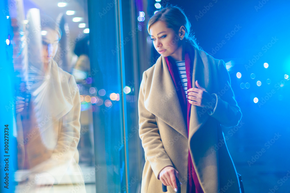 Christmas. A young beautiful woman in the late evening looks at the glowing window and chooses a gift. Colorful, magical lighting, beautiful glare. Cheerful girl wondered at the window, the agony of Christmas. A young beautiful woman in the late evening looks at the glowing window and chooses a gift. Colorful, magical lighting, beautiful glare. Cheerful girl wondered at the window, the agony of