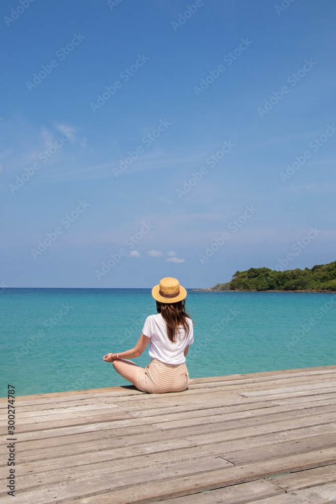 Young woman in a hat sits on a pier near the clear turquoise sea