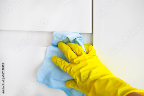 A female hand in yellow rubber gloves wipes a modern white kitchen with a microfiber cloth.