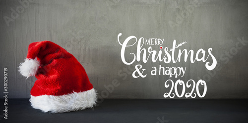 English Calligraphy Merry Christmas And A Happy 2020. Red Santa Claus Hat. Grungy Concrete Background