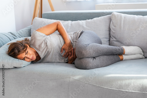 Woman in painful expression holding hands against belly suffering menstrual period pain, lying sad on home bed, having tummy cramp in female health concept photo