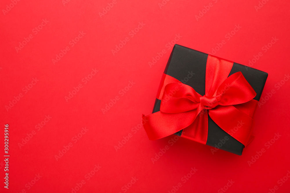 Black gift box with red ribbon and bow on red background.