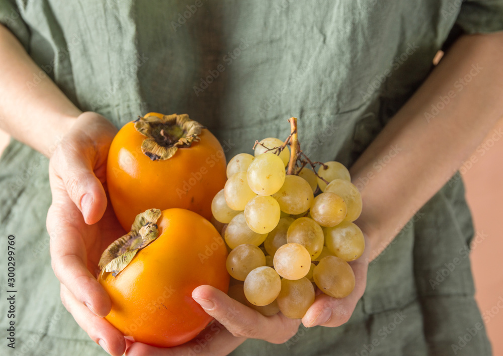 Young Caucasian woman in gray linen shirt holds in hand bunch of ripe juicy white grapes persimmons. Autumn harvest healthy lifestyle plant based diet concept