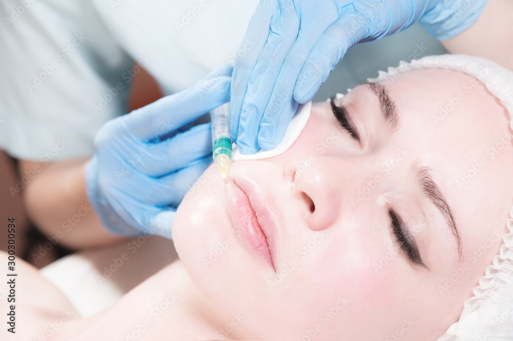 Close-up Attractive young woman gets anti-aging face injections. She lies calmly in a clinic or salon. An experienced young cosmetologist fills female wrinkles with hyaluronic acid from a syringe