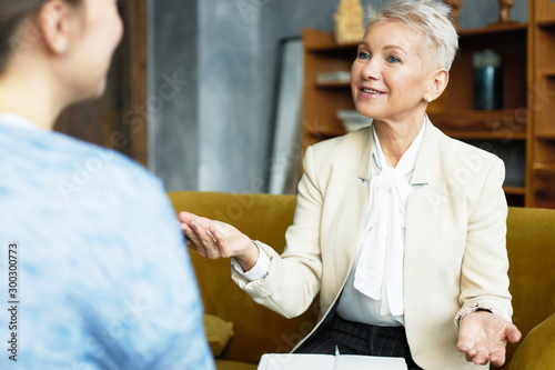 Attractive mature female HR director and job candidate at interview, having lively discussion. Confident middle aged woman in formal wear gesturing emotionally, interviewing applicant in front of her
