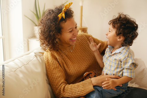Fotomurale Indoor shot of happy young Hispanic woman with brown wavy hair relaxing at home embracing her adorable toddler son