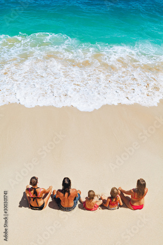 On sunny beach happy kids with parents have fun, sit on white sand see at sea surf with foam and splashes. Top view. Active children lifestyle, summer family vacation travel on tropical family resort.