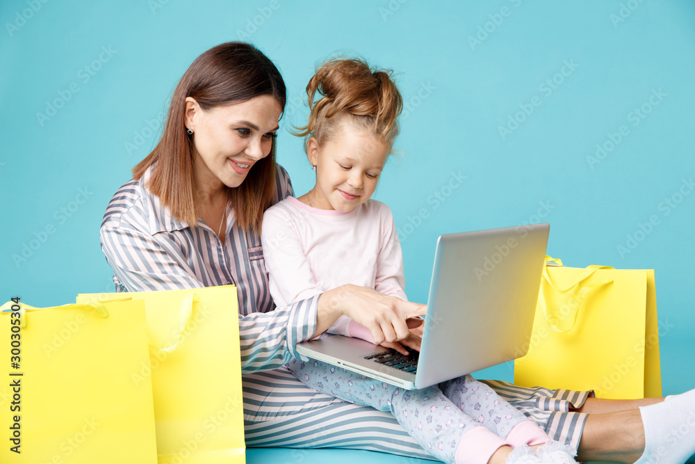 Portrait of mother and daughter with laptop sitting on the floor isolated over the blue studio. Online family shopping concept Portrait of mother and daughter with laptop sitting on the floor isolated over the blue studio. Online family shopping concept