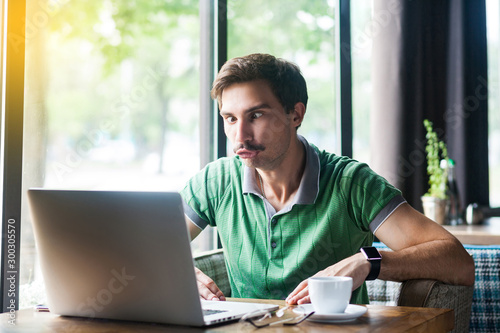 Young funny businessman in green t-shirt sitting and looking at laptop display with stupid crazy face and crossed eyes. business and freelancing concept. indoor shot near big window at daytime. photo