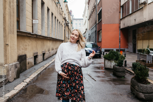 Portrait of beautiful pregnant woman. She is standing in the middle of the street and holding hands on her belly. 