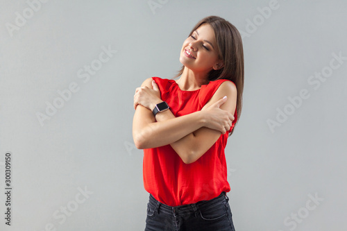 I live myself. Portrait of happy beautiful brunette young woman in red shirt standing, toothy smile and hugging herself and enjoying. indoor, studio shot, isolated on gray background.