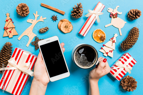 Top view of a woman holding a phone in one hand and a cup of coffee in another hand on blue background. Christmas decorations and toys. New Year holiday concept. Mockup