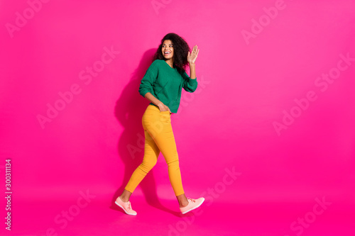 Full length profile side photo of positive curly hair afro american girl had fun on winter free time go walk hold hand say bye wear knitted jumper outfit pants sneakers isolated pink color background