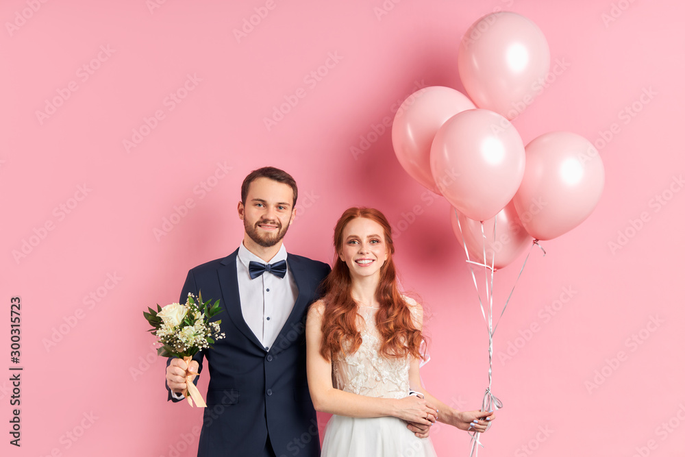 couple in love get married, isolated over pink background happy together, hugging and smiling. Woman holding pink air balloons, man hold wedding bouquet in hands. Look at camera. Wedding, love concept couple in love get married, isolated over pink background happy together, hugging and smiling. Woman holding pink air balloons, man hold wedding bouquet in hands. Look at camera. Wedding, love concept