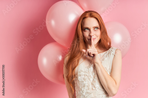Beautiful woman with auburn hair wearing white dress with air balloons behind, thinking, look up. Isolated over pink background