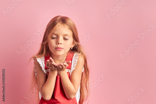 Lovely girl of caucasian ethnicity dressed in red overalls blowing kiss isolated over pink background