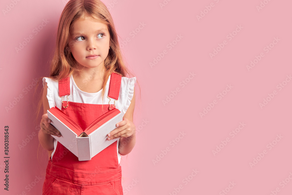 Thoughtful caucasian girl in red overalls holding white book and dream looking side, isolated over pink background Thoughtful caucasian girl in red overalls holding white book and dream looking side, isolated over pink background