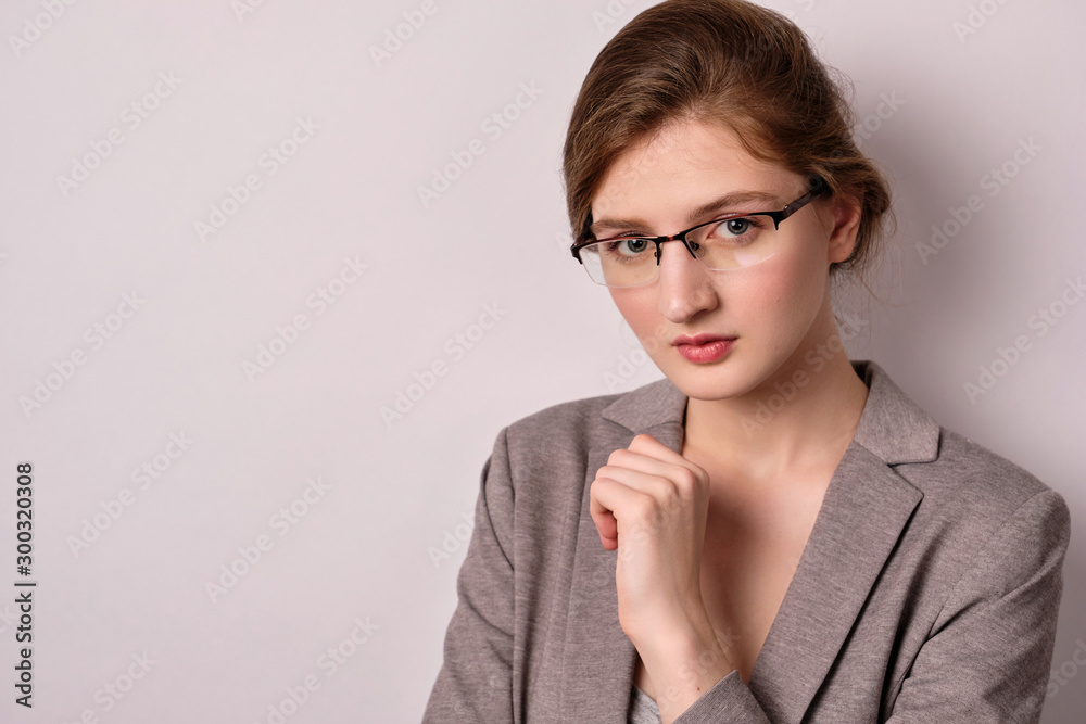 A girl in a gray jacket and glasses stands on a light background and looks thoughtfully into the frame. A girl in a gray jacket and glasses stands on a light background and looks thoughtfully into the frame.