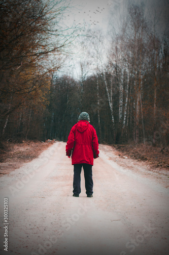 Photo of a man in the back. A man in a red jacket stands on a road surrounded by forest. A person is traveling. Season autumn, winter or early spring. The background is blurred.