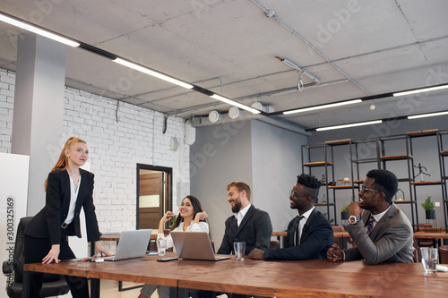 Diverse smiling people of different nations gathered at conference table to discuss business ideas with caucasian female boss with auburn hair, everyone looking at her. Office background