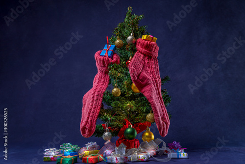 There is Christmas tree with different colors balls and pink wool present socks. There are different colors gift with ribbons and bows near the tree. Merry Christmas. Happy New Year 2020. 