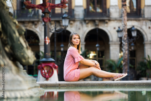Attractive girl tourist in dress sitting at stone bench near fountain in Barcelona
