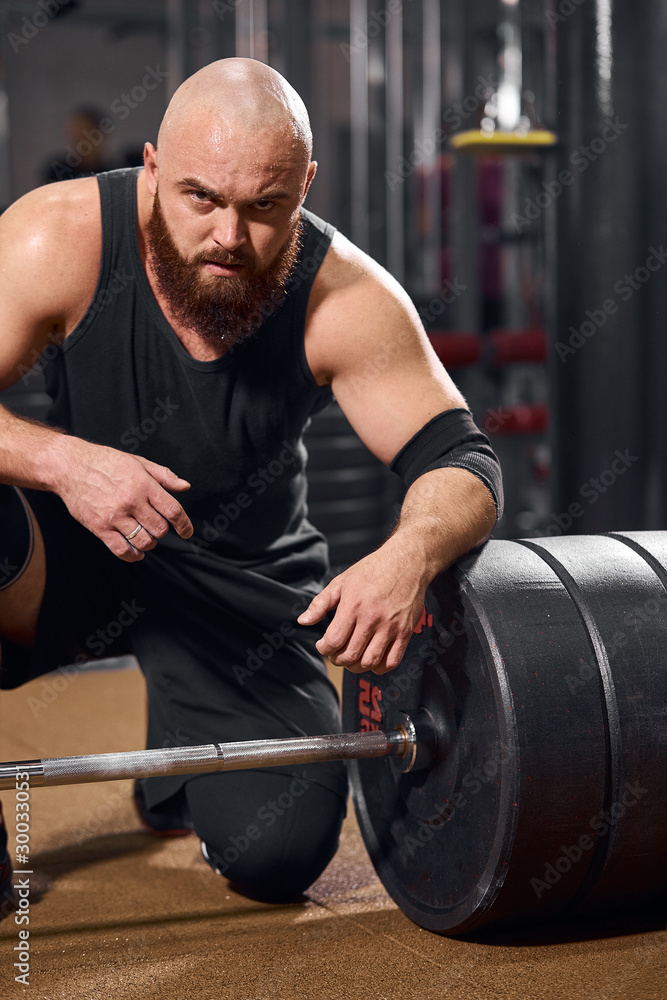 Portrait of brutal powerful bodybuilder sitting near heavy barbell, putting knee on floor, looking strongly at camera, posing in modern fitness studio