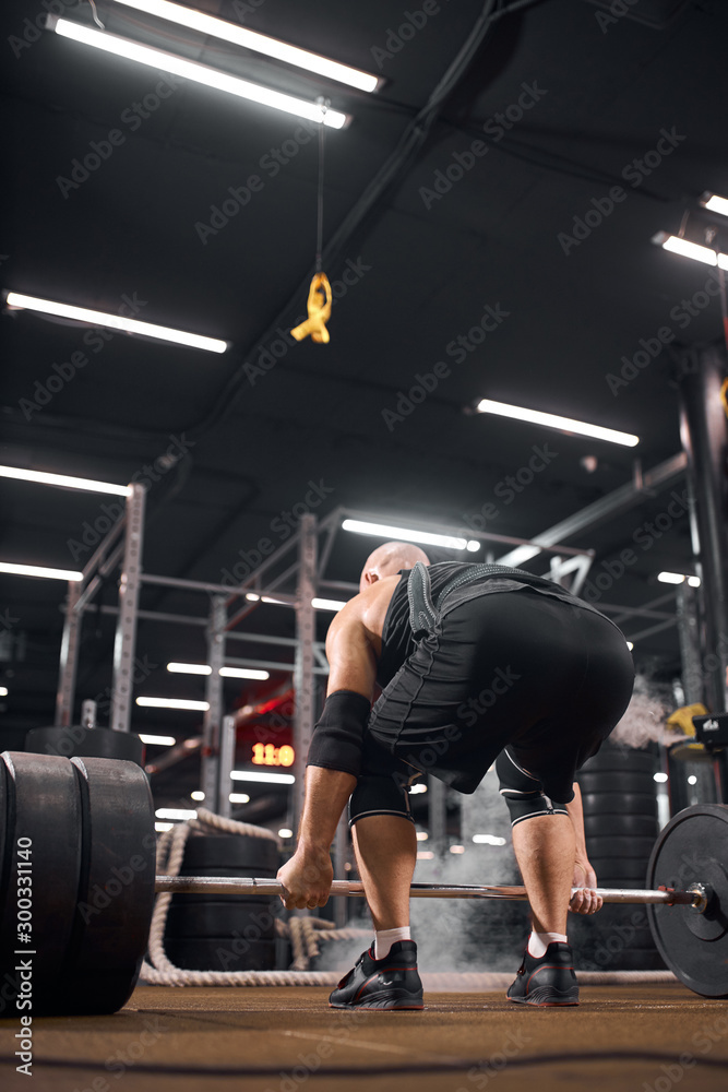 Bottom shot of strong brutal powerlifter dressed in black sportswear, bending in front of heavy barbell, standing with his back towards camera, modern fitness center concept, indoor shot
