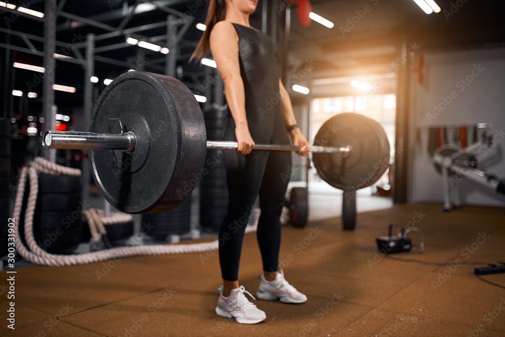 Young unrecognizable woman with heavy barbell in hands, doing squats, preparing for powerlifting competition, spending time in modern gym, active people concept, side sho Young unrecognizable woman with heavy barbell in hands, doing squats, preparing for powerlifting competition, spending time in modern gym, active people concept, side sho