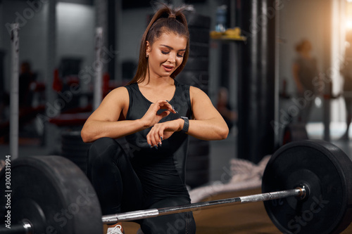 Attractive sporty female athlete happily looking at watch happy to finish training, sitting on floor near heavy barbell, spending time in professional fitness studio, portrait, close view