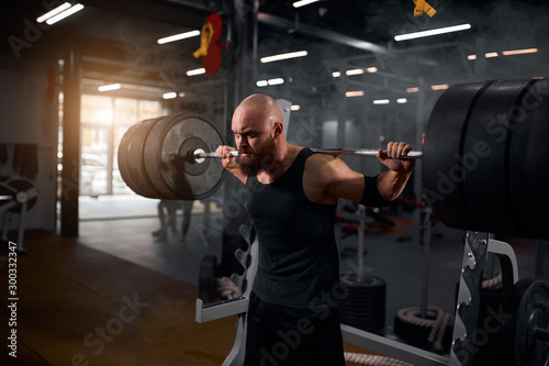 Strong active hairless athlete with thick brunette hard trying to rise hands with heavy barbell row above head, looking away with severe look, training in underground garage, black tone, portrait,