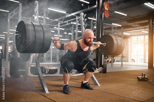 Fototapeta Naklejka Na Ścianę i Meble -  Acctive strong weightlifter sitting with heavy barbell in hands, squatting in modern gym room, professional sport concept, white smoke in the air, indoor side shot