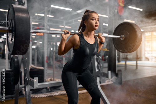 Portrait of sporty young fit woman spending time in modern fitness studio  pumping up muscles  using heavy barbell  preparing for workout at worldwide competition of bodybuilding