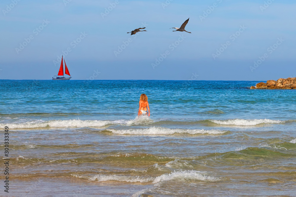 Young woman in a swimsuit goes in the surf. Sailboat with a red sail on the horizon. Sunny calm sea seascape. Migrating birds flying in the sky