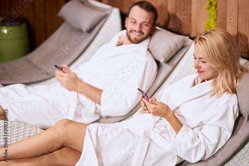 Cheerful handsome boyfriend looking at blonde young woman sitting in comfortable armchair, holding cell phones in hands, looking away with positive mood, good feelings after spa procedure