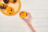cropped view of woman taking delicious cupcake with spiders from orange plate on white wooden table, Halloween treat