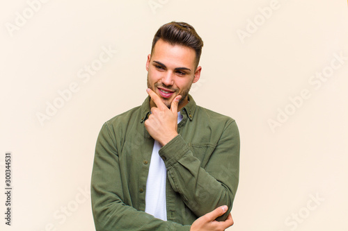 young hispanic man smiling with a happy, confident expression with hand on chin, wondering and looking to the side against isolated background photo