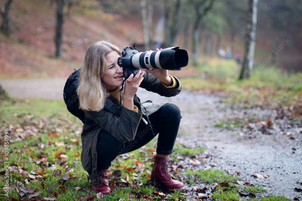 Blonde photographer squatting and using camera with large telephoto lens while taking shots in nature - obrazy, fototapety, plakaty 