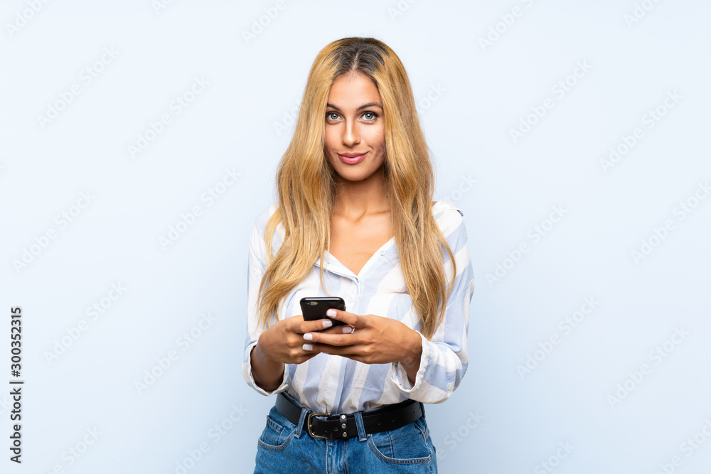 Young blonde woman over isolated blue background sending a message with the mobile Young blonde woman over isolated blue background sending a message with the mobile