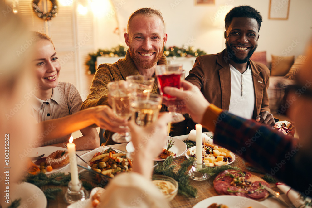 Multi-ethnic group of cheerful adult people clinking champagne glasses while enjoying Christmas dinner at home, copy space