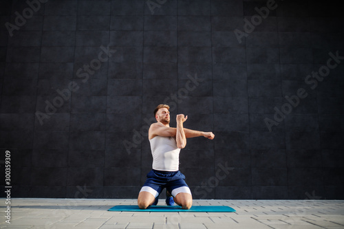 Handsome muscular bearded caucasian man kneeling on mat outdoors and stretching his arms. In background is gray wall.