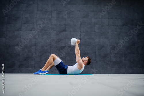Side view of handsome caucasian muscular man lying down on mat and lifting kettle bell.