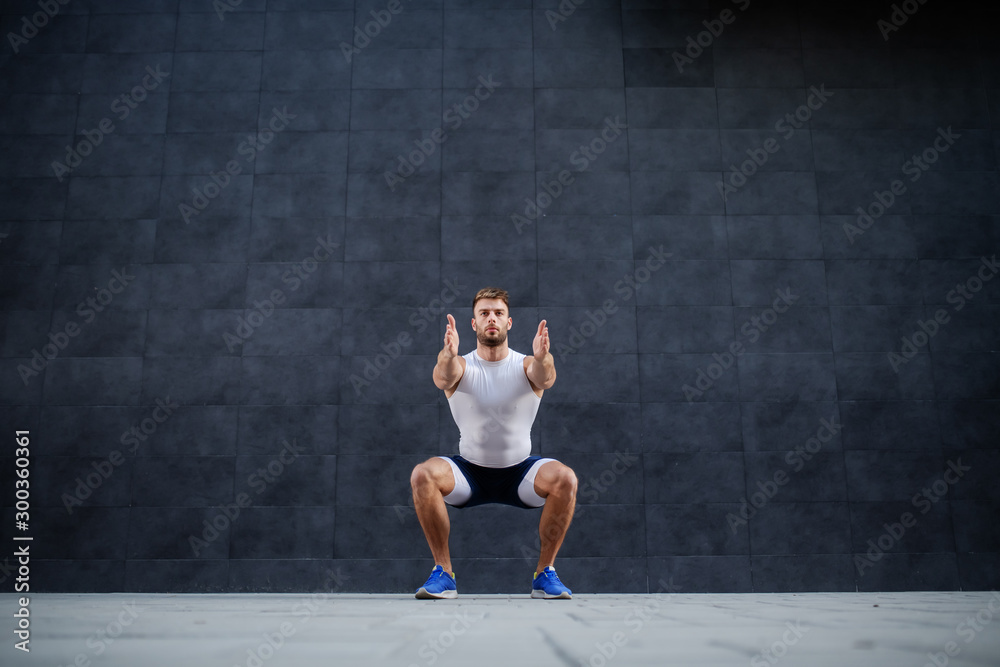 Handsome muscular caucasian man in shorts and t-shirt doing squatting exercise outdoors. In background is gray wall. Handsome muscular caucasian man in shorts and t-shirt doing squatting exercise outdoors. In background is gray wall.