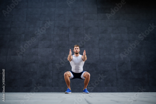 Handsome muscular caucasian man in shorts and t-shirt doing squatting exercise outdoors. In background is gray wall.