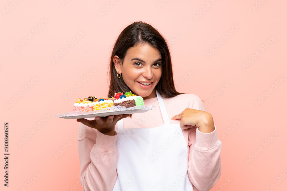 Young girl holding lots of different mini cakes over isolated background with surprise facial expression Young girl holding lots of different mini cakes over isolated background with surprise facial expression